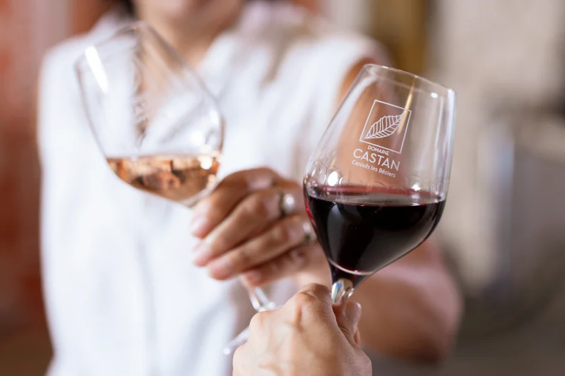 Plan rapproché de dégustation : deux verres siglés 'Domaine Castan - Cazouls les Béziers' tenus par des mains, l'un rempli de rosé, l'autre de rouge, lumière douce. Photo professionnelle élégante qui met en valeur l'expérience de dégustation et l'image de marque. Excellente pour une page caveau/dégu