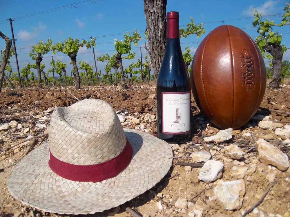 Mise en scene lifestyle : une bouteille de rouge Domaine Chemin de Notre Dame, un chapeau de paille et un ballon de rugby en cuir vintage poses sur le sol caillouteux du vignoble, vignes et ciel bleu en arriere-plan. Resume l'identite du domaine (rugby + vigne).