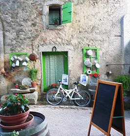 Coin de cour du domaine: facade en pierre ancienne, volets verts, velo blanc appuye, pots de fleurs, assiettes decoratives et ardoise. Ambiance pittoresque et accueillante du lieu (gite/oenotourisme). Image petite (266x280) mais charmante.