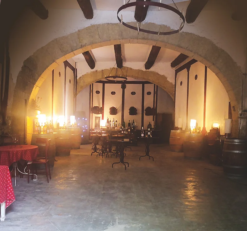 Caveau/chai voute en pierre ancienne, grandes arches, poutres apparentes, lustres en fer forge, barriques et tables de degustation (nappe rouge) baignees d une lumiere chaude. Tres beau cadre patrimonial pour la degustation. Resolution 800px.