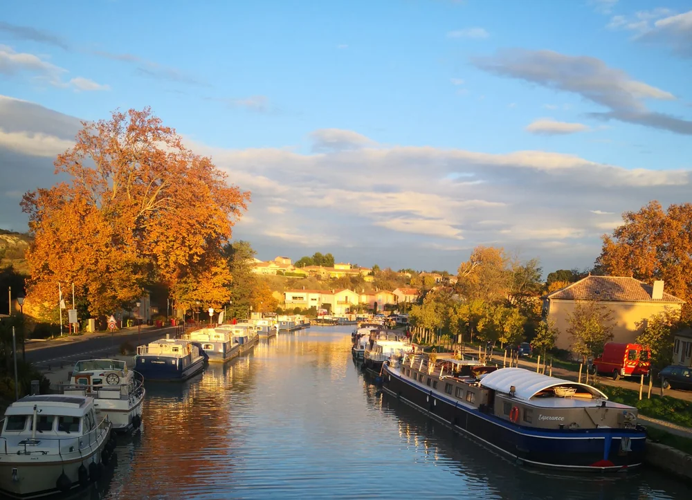 Le Canal du Midi a Capestang en automne: peniches et bateaux amarres, platanes aux feuillages orange, village et reflets dans l eau sous une lumiere doree. Beau paysage de region qui ancre le domaine dans son environnement touristique (Canal du Midi). Grande resolution.