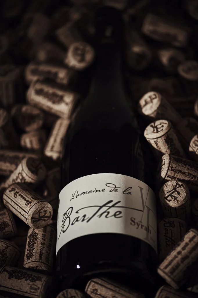 Photo d'ambiance: bouteille Domaine de la Barthe Syrah nichee dans un lit de bouchons de liege marques au nom du domaine, eclairage sombre et dramatique. Tres soigne, mise en valeur premium.