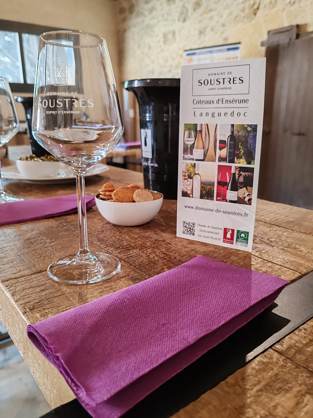 Verre grave au logo du domaine et depliant 'Coteaux d'Ensérune' poses sur une table en bois avec petite coupelle de biscuits apero et serviette violette. Mise en situation de la degustation au caveau (format vertical).