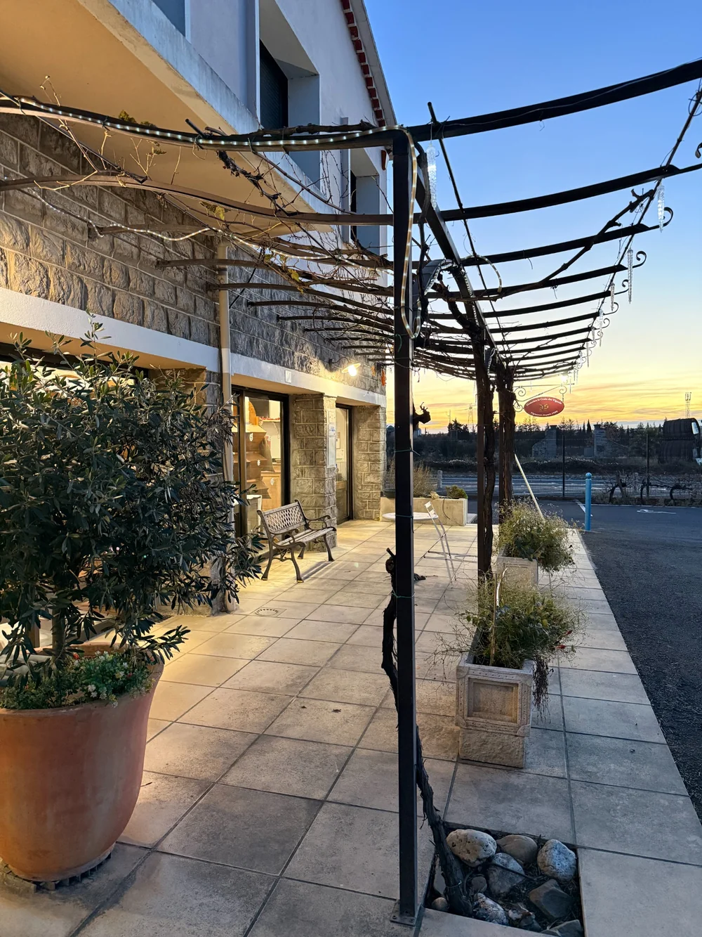 Vue laterale de la terrasse du caveau au crepuscule, treille et plantes en pot, ciel orange. Doublon d'ambiance de la facade.