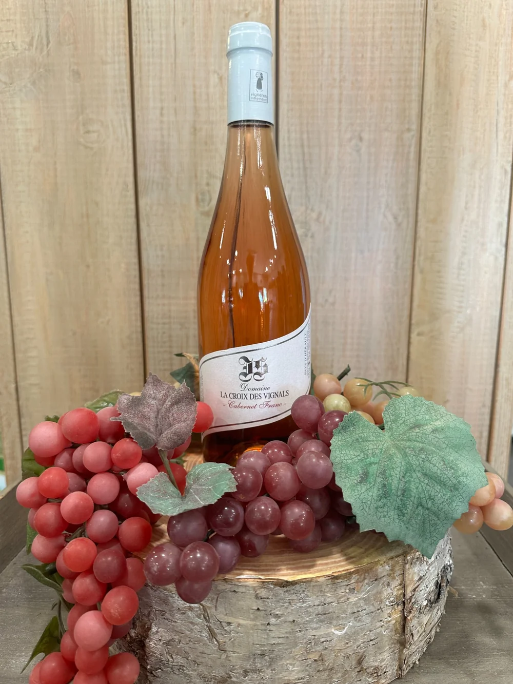 Packshot lifestyle d'une bouteille de rose La Croix des Vignals (Cabernet Franc) posee sur une bille de bois, entouree de grappes rouges et de feuilles de vigne, fond bois. Composition soignee.