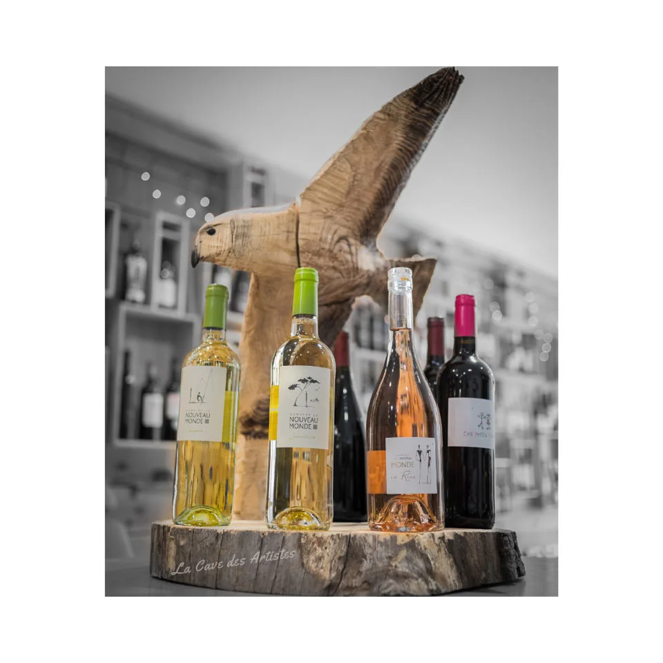 Mise en scene lifestyle de la gamme du domaine (blancs, rose, rouges) disposee sur une rondelle de bois avec une sculpture en bois d'un oiseau de proie, fond de cave/boutique desature. Belle photo de bouteilles en situation, pas une etiquette plate.