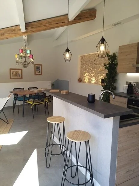 Interieur lumineux du gite du domaine : cuisine ouverte et coin repas sous charpente apparente, mur de pierre, suspensions design, lustre colore. Soigne et accueillant. 463x617.