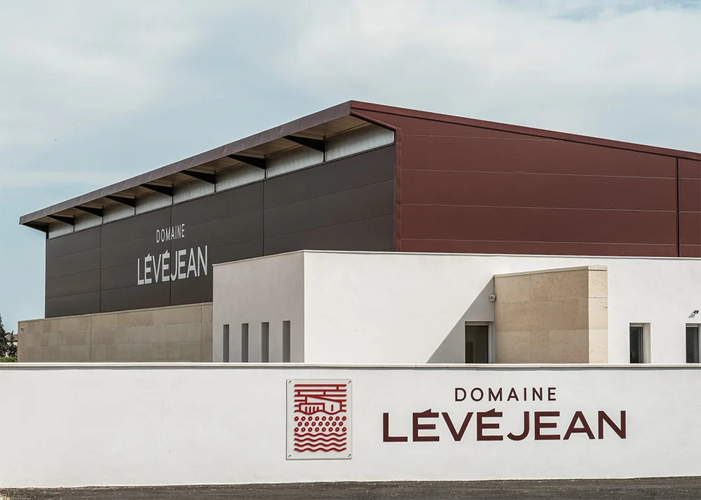Architecture du domaine : le batiment moderne du chai (bardage bordeaux et anthracite, enseigne DOMAINE LEVEJEAN) derriere un muret blanc portant le logo grave. Image nette et soignee de l'identite architecturale. 1024x732.