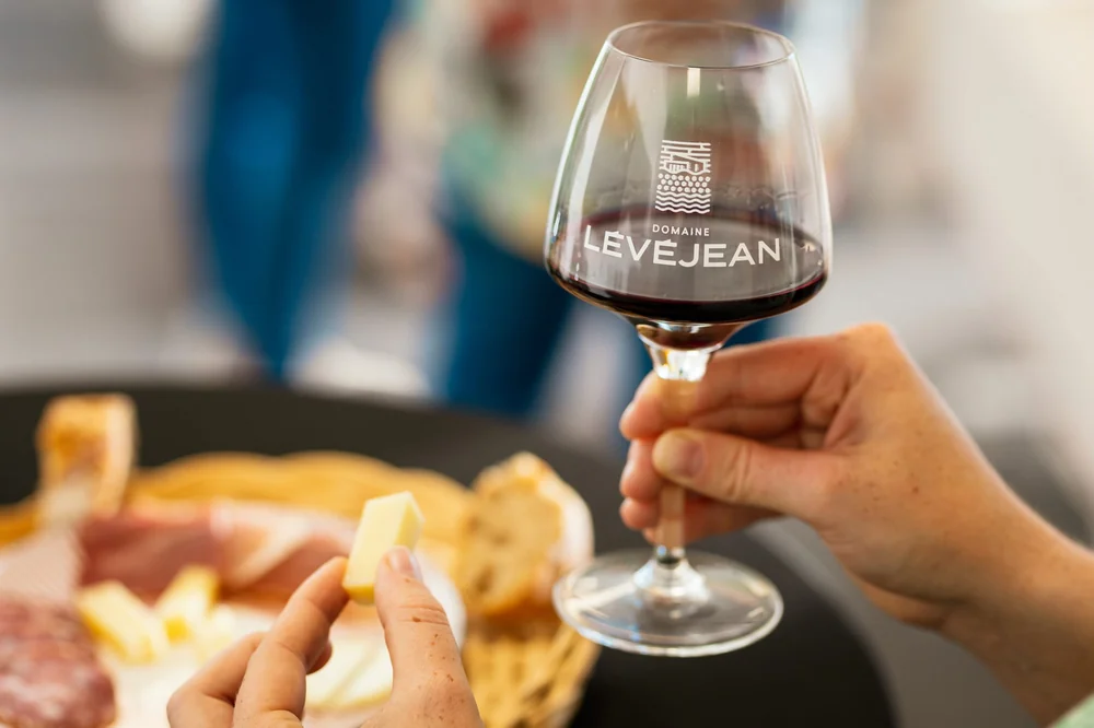 Cliche lifestyle professionnel : une main tient un verre logote Domaine Levejean rempli de vin rouge, avec en avant-plan flou une planche de charcuterie/fromage et du pain. Mise en situation chaleureuse (pas une etiquette plate). Photo Herve Leclair, 2560x1707.