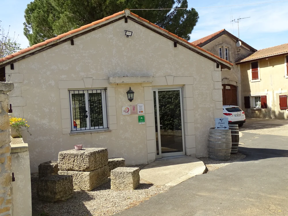 Extérieur du domaine : façade en pierre du caveau/boutique avec toiture en tuiles romanes, applique en fer forgé, plaques de certifications près de la porte vitrée, vieux fûts décoratifs et blocs de pierre dans la cour ensoleillée, bâtiments du mas en arrière-plan. Donne à voir l'arrivée et l'archit