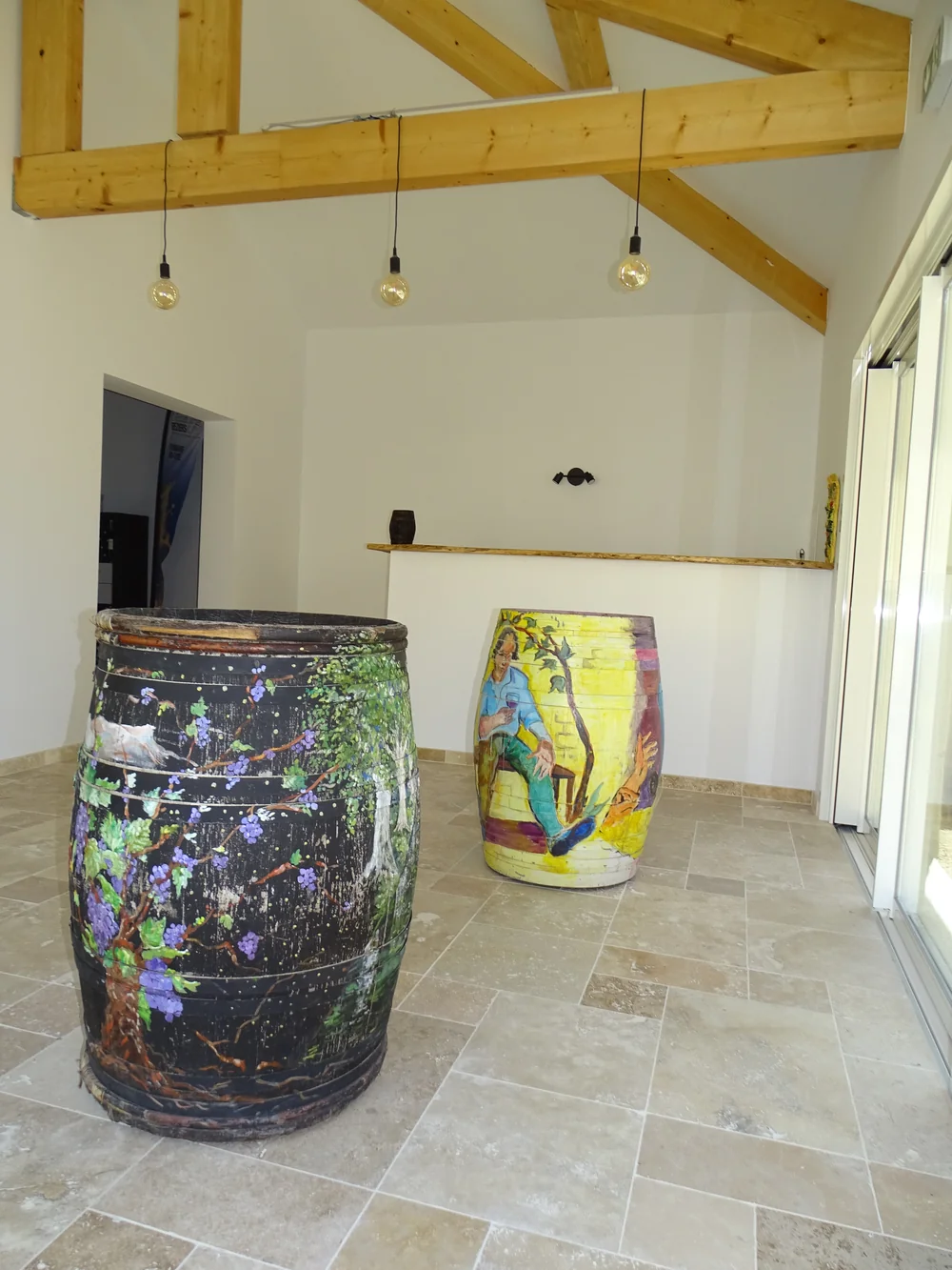 Salle de dégustation / caveau lumineux : charpente bois apparente, ampoules à filament suspendues, sol en pierre, et deux grands fûts décoratifs peints à la main (l'un orné de pampres et grappes de raisin, l'autre d'un personnage dégustant un verre de vin). Ambiance chaleureuse et soignée, très valo