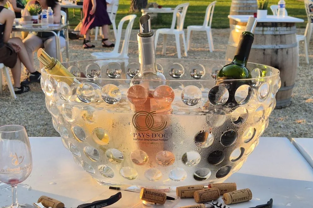 Mise en situation lifestyle (1050x700) : trois bouteilles (blanc, rose, rouge) rafraichissant dans une grande vasque a glacons transparente lors d'une degustation au jardin a l'heure doree, fut et verre en arriere-plan. Belle ambiance, met les vins en valeur (pas une etiquette plate).