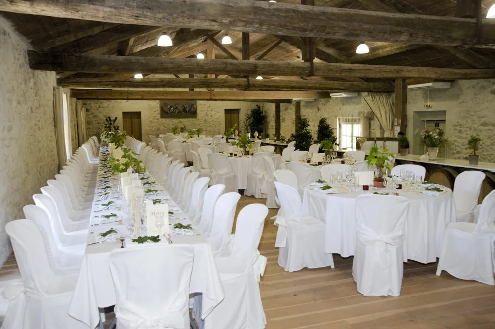 Salle de reception / caveau (1500x998) sous belle charpente de poutres anciennes et murs de pierre, dressee pour un repas (nappes blanches, tables rondes et grande tablee), avec des futs de chene en decor. Montre le caractere du lieu evenementiel et le chai.