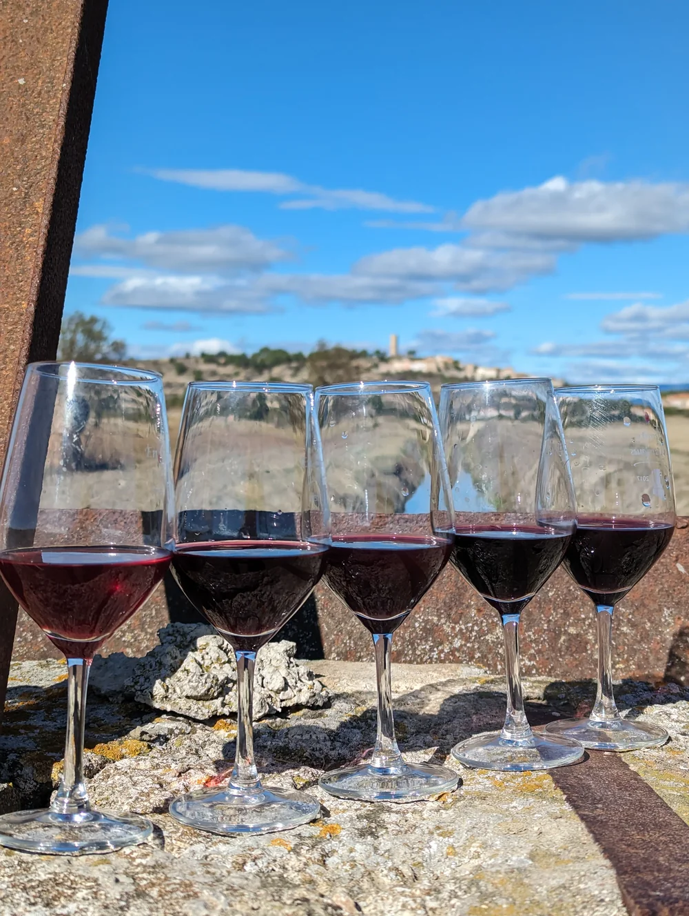Cinq verres de vin rouge alignes sur un muret de pierre, avec en arriere-plan le paysage de l'etang et une tour (Oppidum d'Enserune) sous un ciel bleu nuageux. Belle association vin + terroir, lifestyle.