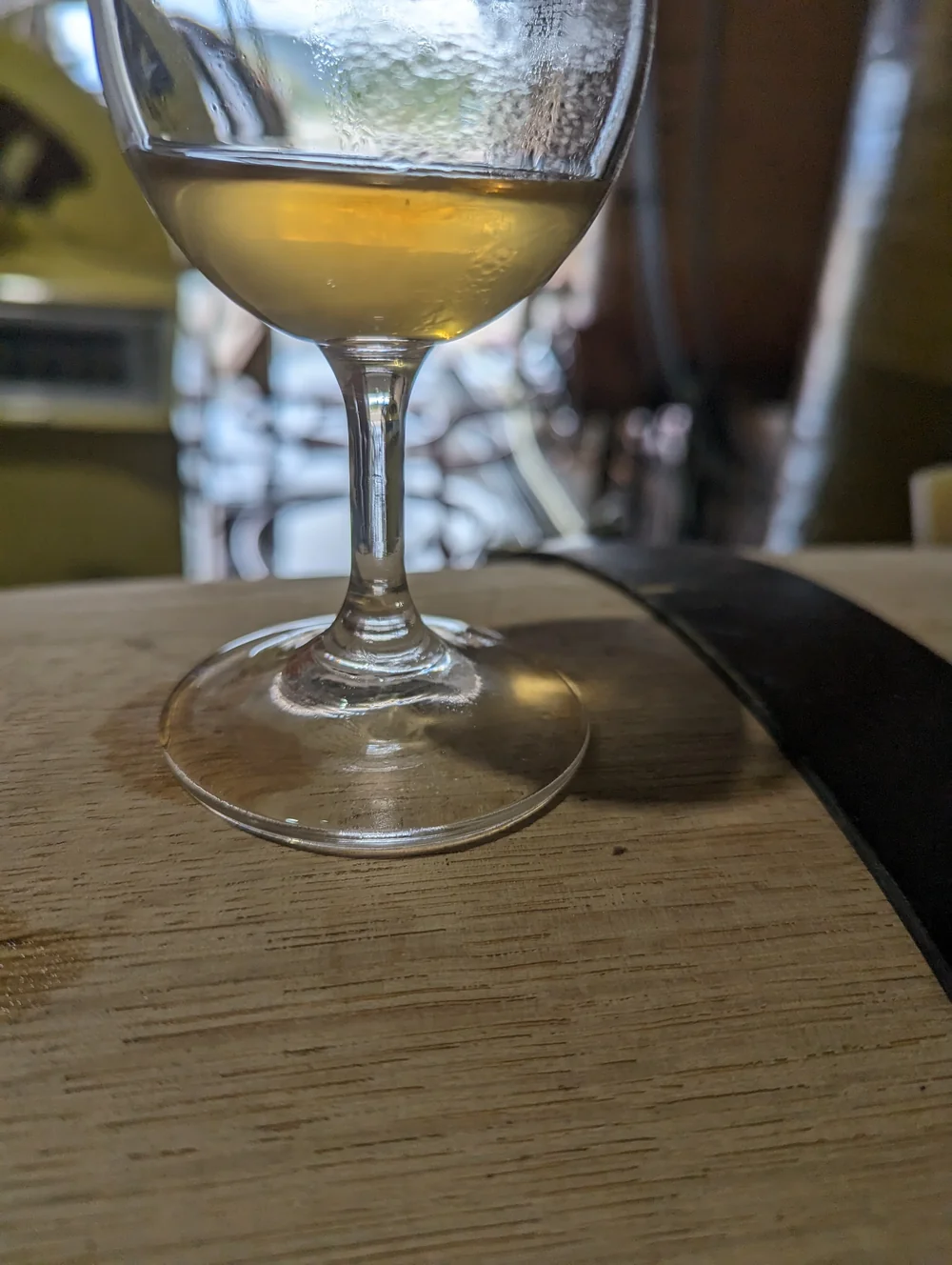 Verre de vin blanc pose sur la tete d'une barrique dans le chai, faible profondeur de champ, cuves en arriere-plan flou. Cliche de degustation/elevage intime.