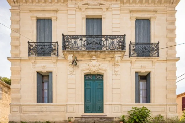 Belle façade en pierre du mas/bâtiment du domaine : fenêtres à balcons en fer forgé, porte ancienne, fronton sculpté. Architecture languedocienne authentique, met en valeur le lieu.