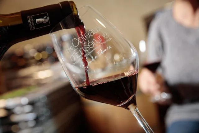 Packshot lifestyle : verre de vin rouge gravé 'Coteaux d'Ensérune' tenu en main, bouteille floue à l'arrière, ambiance caveau. Belle photo d'ambiance de dégustation, pas une étiquette plate.