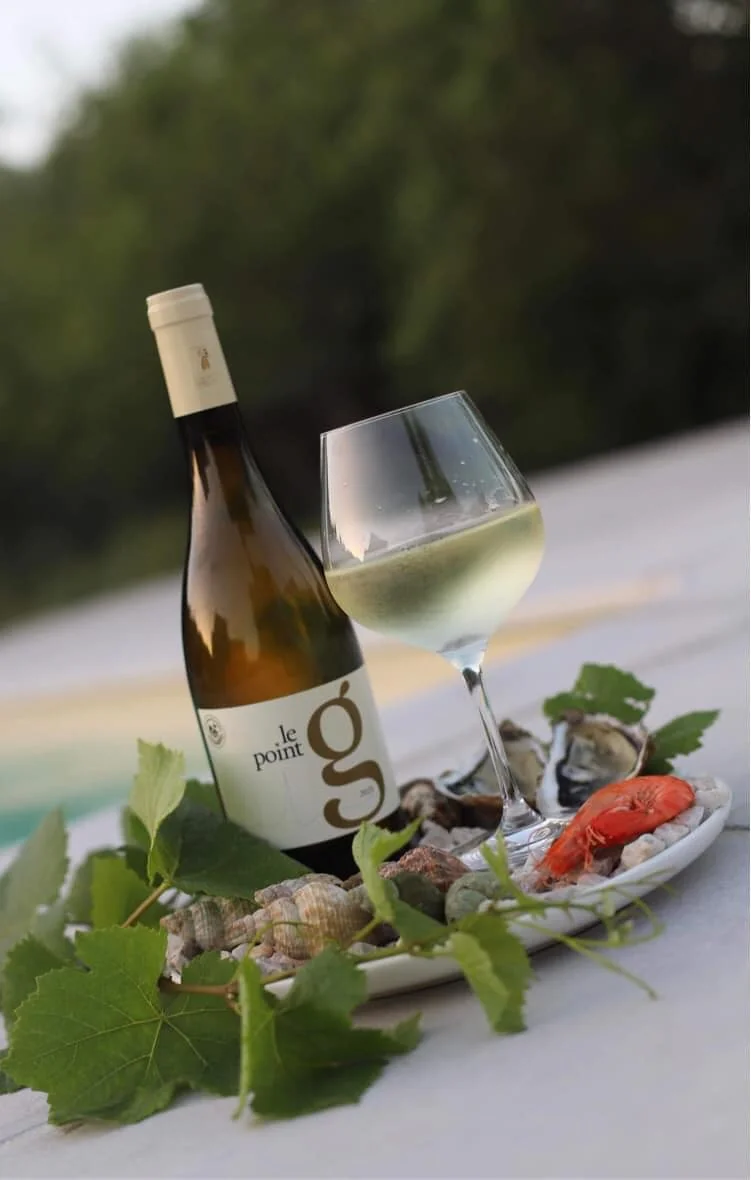 Packshot lifestyle : bouteille de cuvee Le Point G blanc avec un verre de vin blanc et un plateau de fruits de mer (huitres, gambas, coquillages) decore de pampres de vigne, au bord d'une piscine, vegetation floue en arriere-plan. Mise en situation gourmande et estivale, oenotourisme.