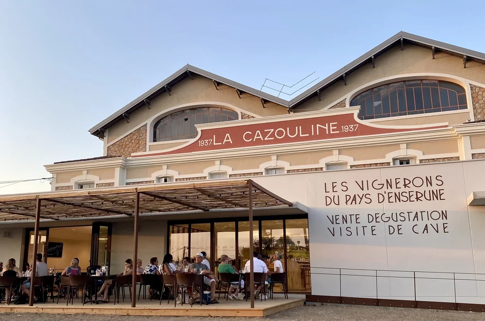 Facade du caveau 'La Cazouline 1937 - Les Vignerons du Pays d'Enserune' au coucher du soleil, avec une terrasse animee remplie de clients attables. Lumiere doree, tres accueillant et qualitatif. Excellent visuel de presentation du lieu.