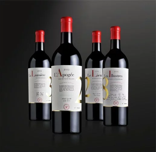 Packshot studio de quatre bouteilles de cuvees prestige (La Lumiere, l'Apogee Saint-Chinian, Le Lien, Les Illustres) sur fond degrade noir. Mise en scene soignee, pas une simple etiquette plate. Resolution moyenne (516px).