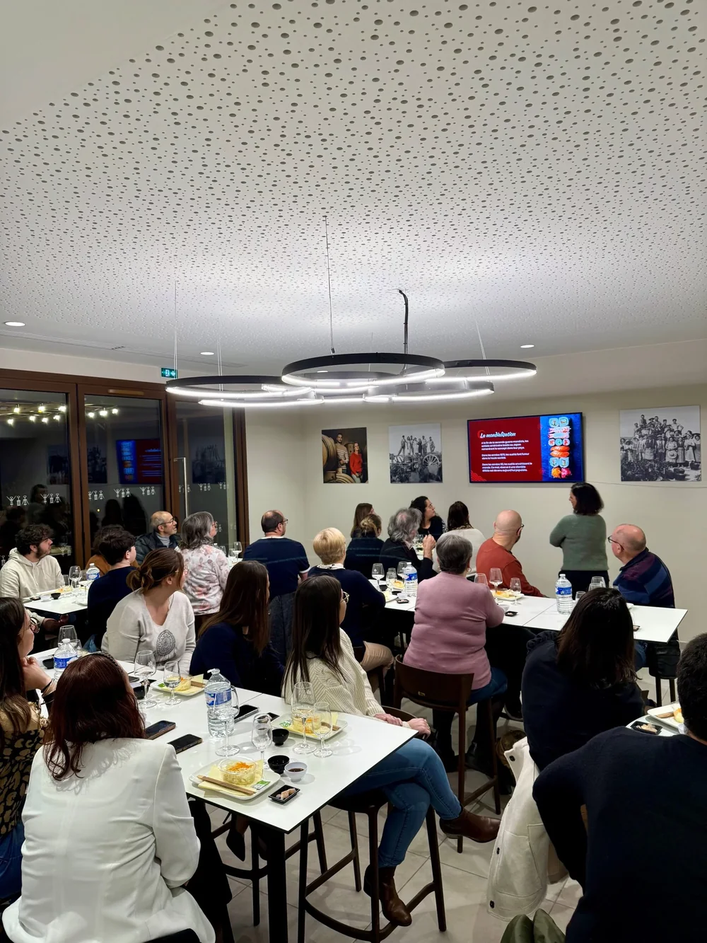 Atelier/soiree de degustation dans la salle renovee : nombreux participants attables avec verres et planches, suspension design et photos historiques au mur. Montre l'activite oenotouristique et la convivialite. Qualite reportage (un peu sombre).