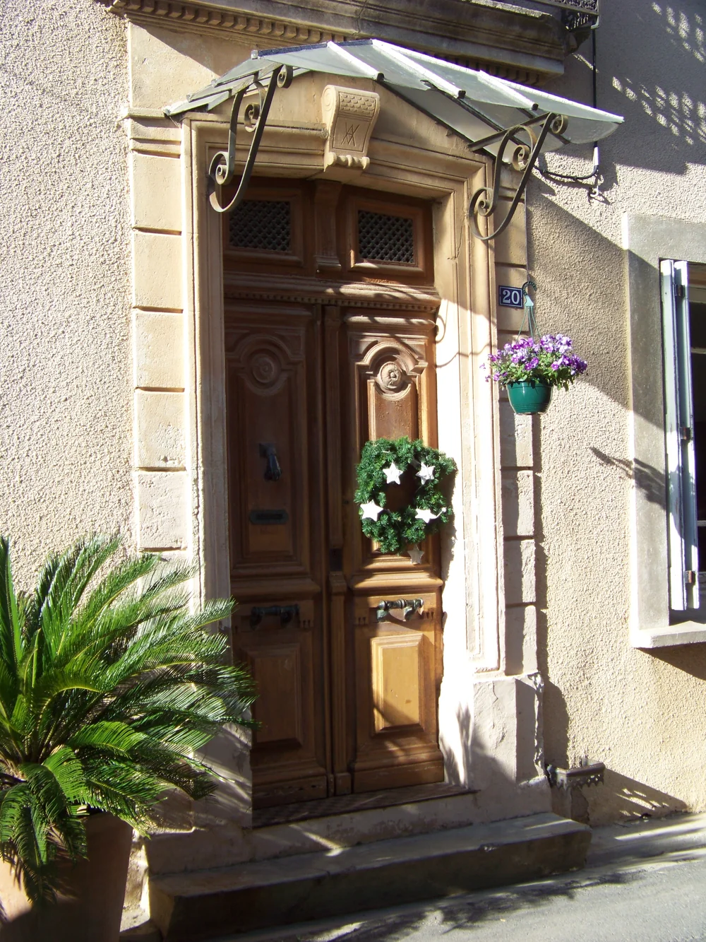 Porte d'entree du domaine au 20 rue Voltaire (adresse confirmee) : belle porte en bois sculpte sous un auvent en ferronnerie, couronne decorative, jardiniere de fleurs violettes et palmier en pot. Facade en pierre baignee de lumiere. Image chaleureuse et authentique de l'entree du mas/maison de vill