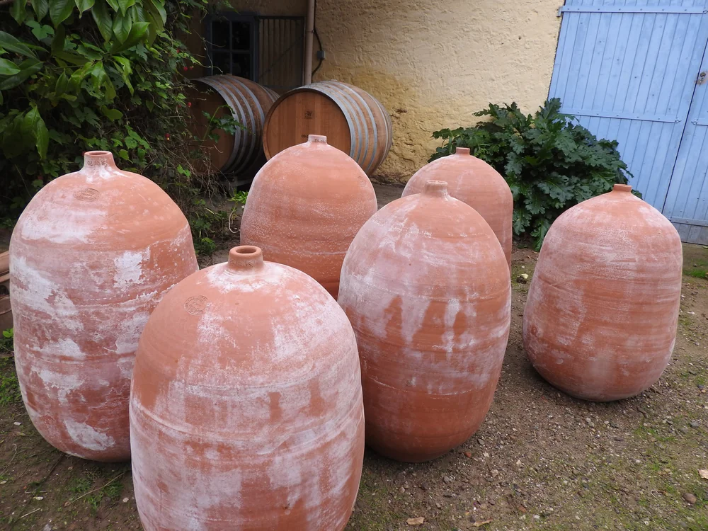 Six grandes amphores en terre cuite (jarres de vinification) alignees dans une cour, devant des barriques de chene et une porte bleue lavee par le temps. Illustre directement la vinification en amphores revendiquee par le domaine. Tres caracteristique et identitaire.